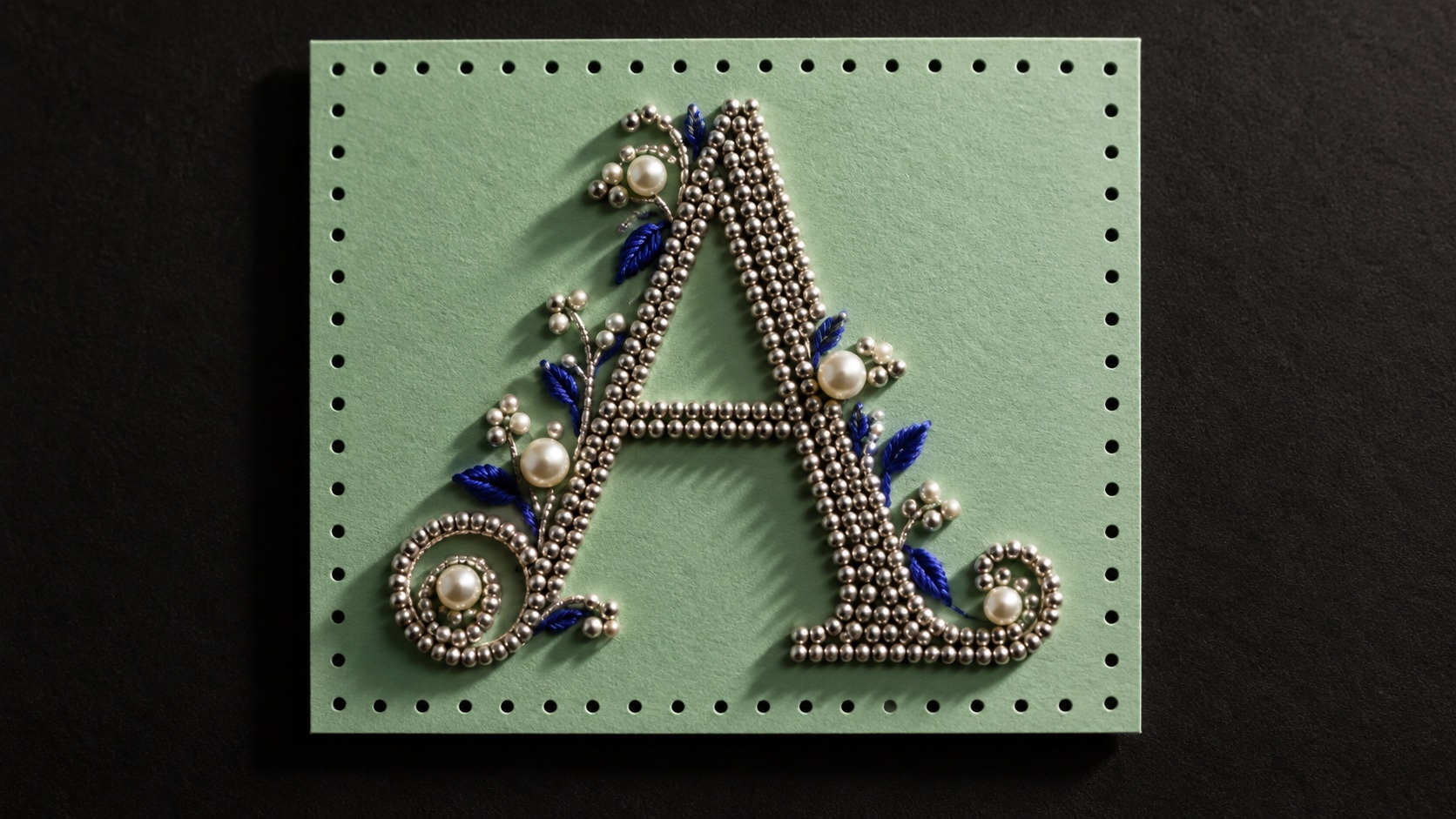 A capital A in chrome beads with pearls and cobalt stones on a mint card with stitched holes around its edge.
