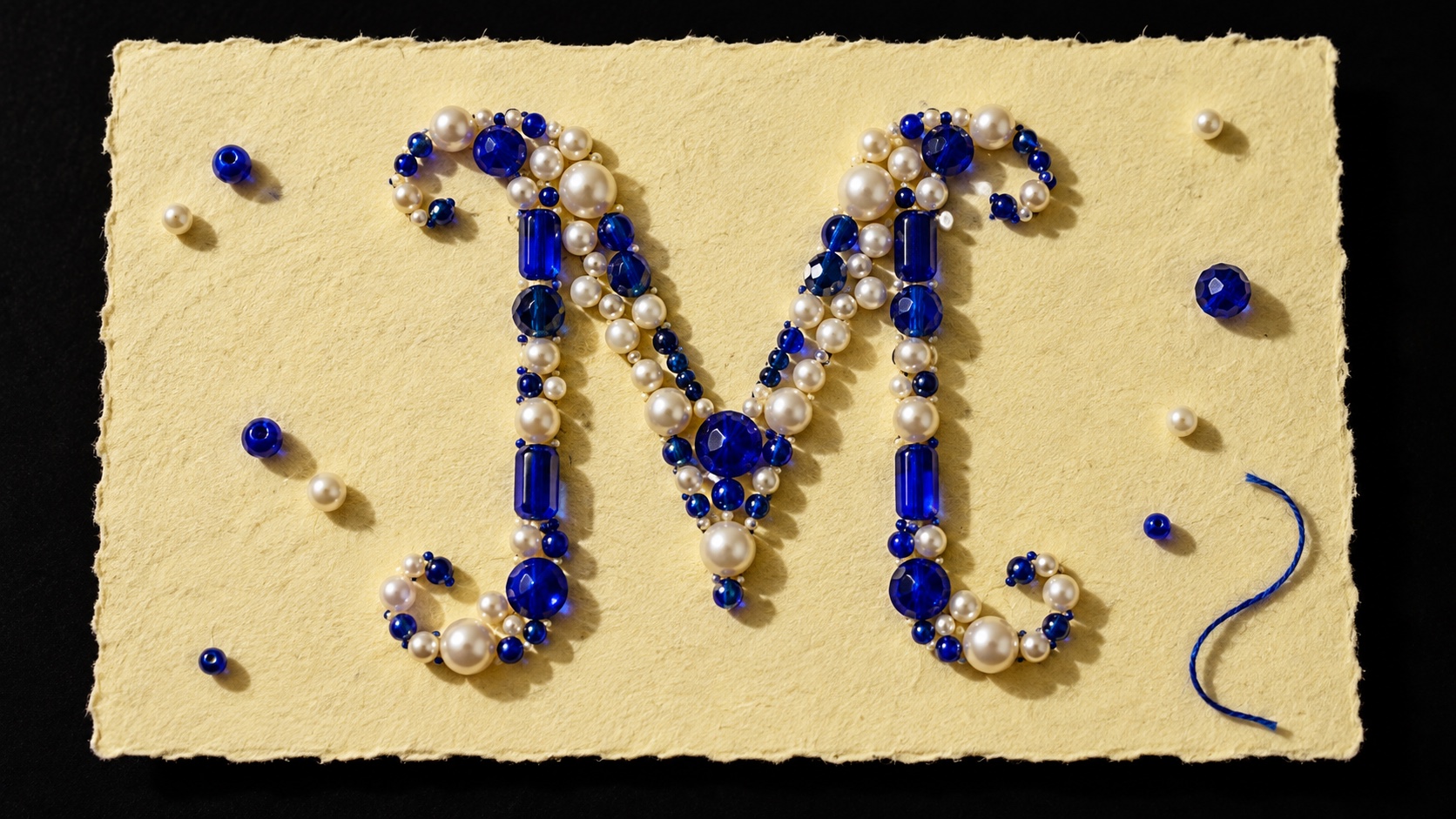 A script M in cobalt stones, pearls and chrome beads on torn cream paper, a loose blue thread curling beside it.