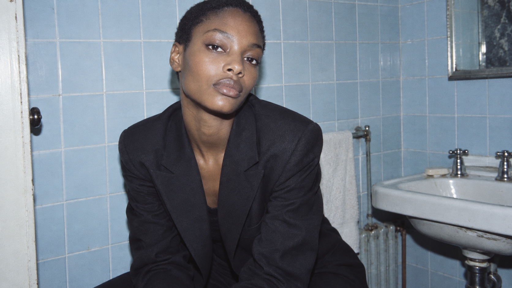 A Black woman in a dark blazer is photographed with hard flash against blue tiles.