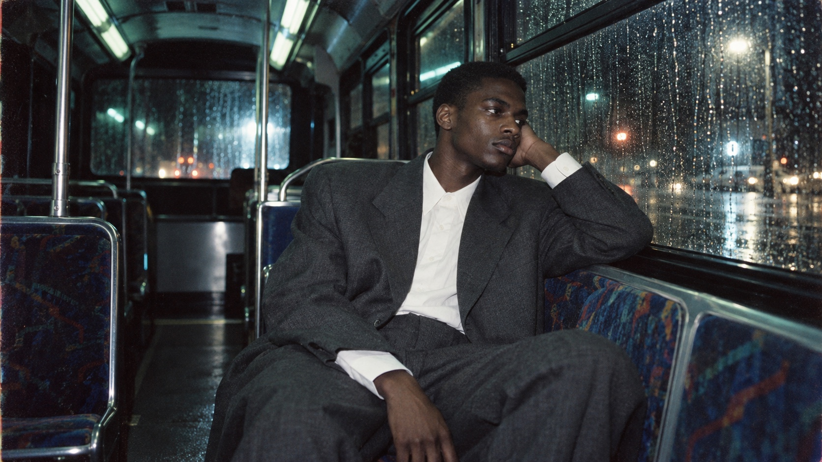 A Black man in a charcoal suit sits alone on a rain-streaked bus at night.