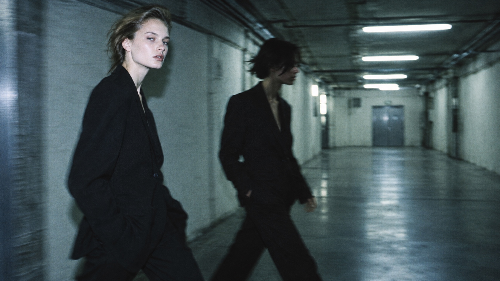 Two white models in black suits cross a fluorescent industrial passage in motion blur.