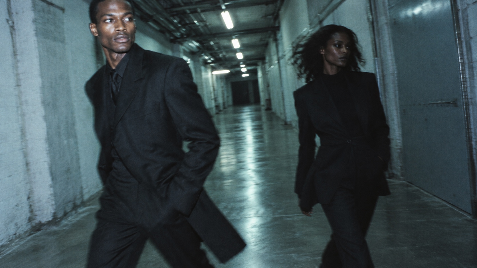 Two Black models in oversized black suits move through a blue-green industrial corridor.