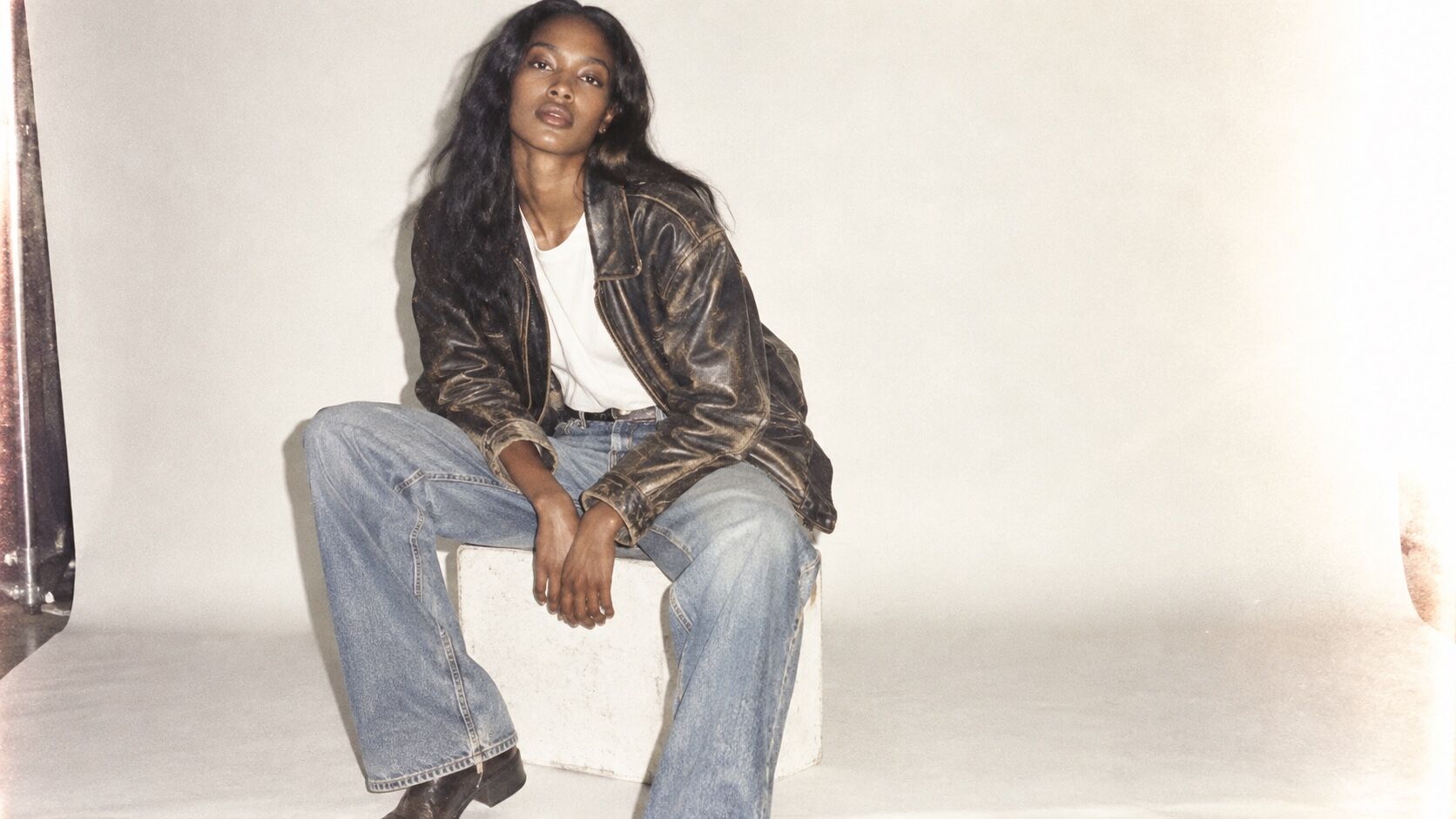 A Black woman in worn leather and pale denim sits against a washed-out studio sweep.
