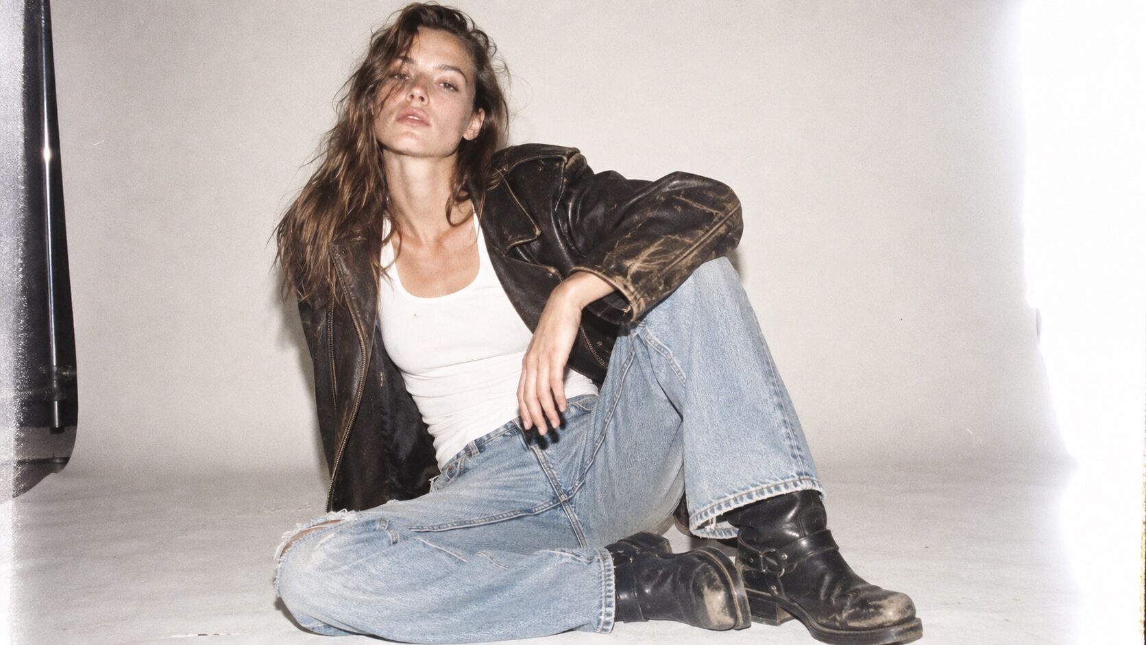 A white woman in worn leather and pale denim sits in a stark flash-lit studio.