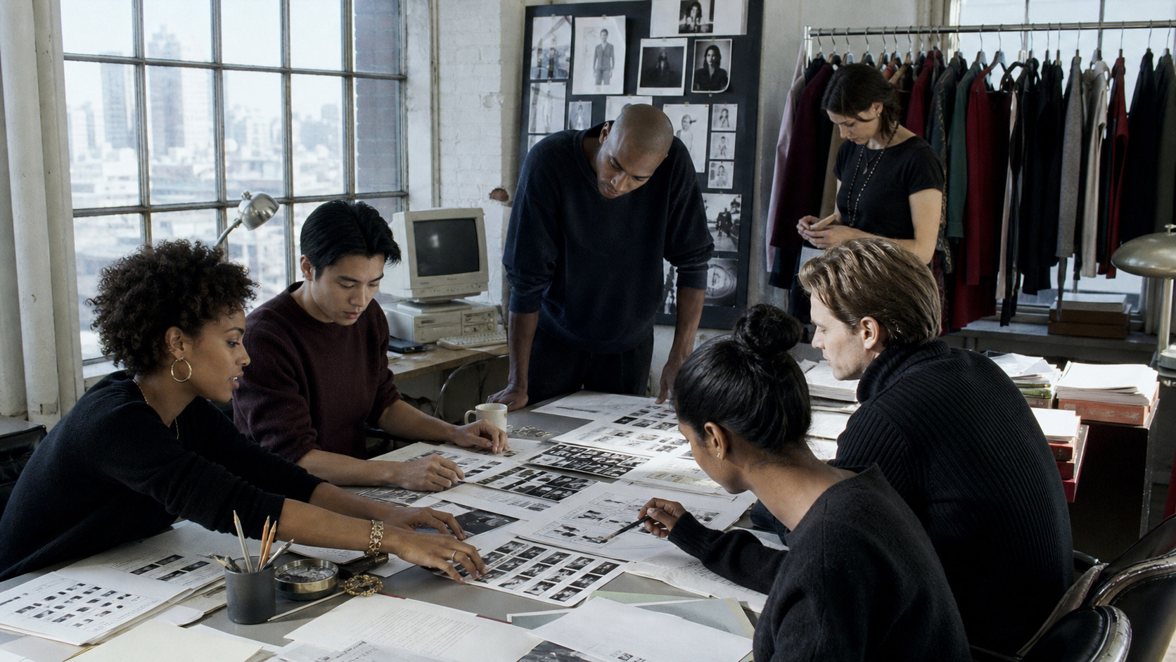 A diverse editorial team collaborates around fashion layouts in a daylight studio.