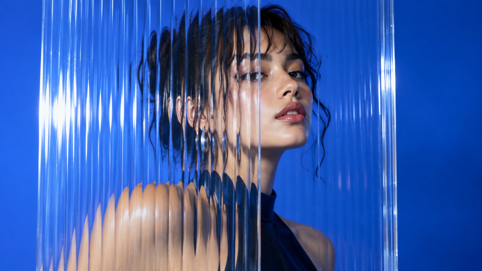 A woman with dark hair seen through vertical fluted glass against a saturated cobalt ground.