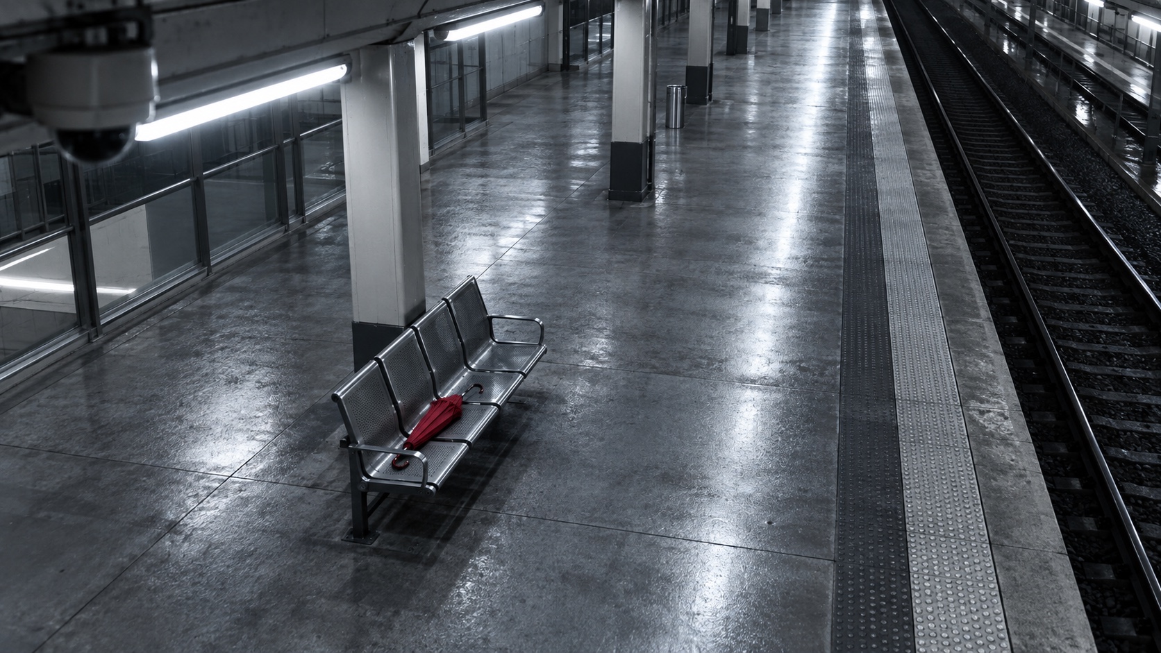 An empty station platform at night with a single red bench, seen from a raised camera.