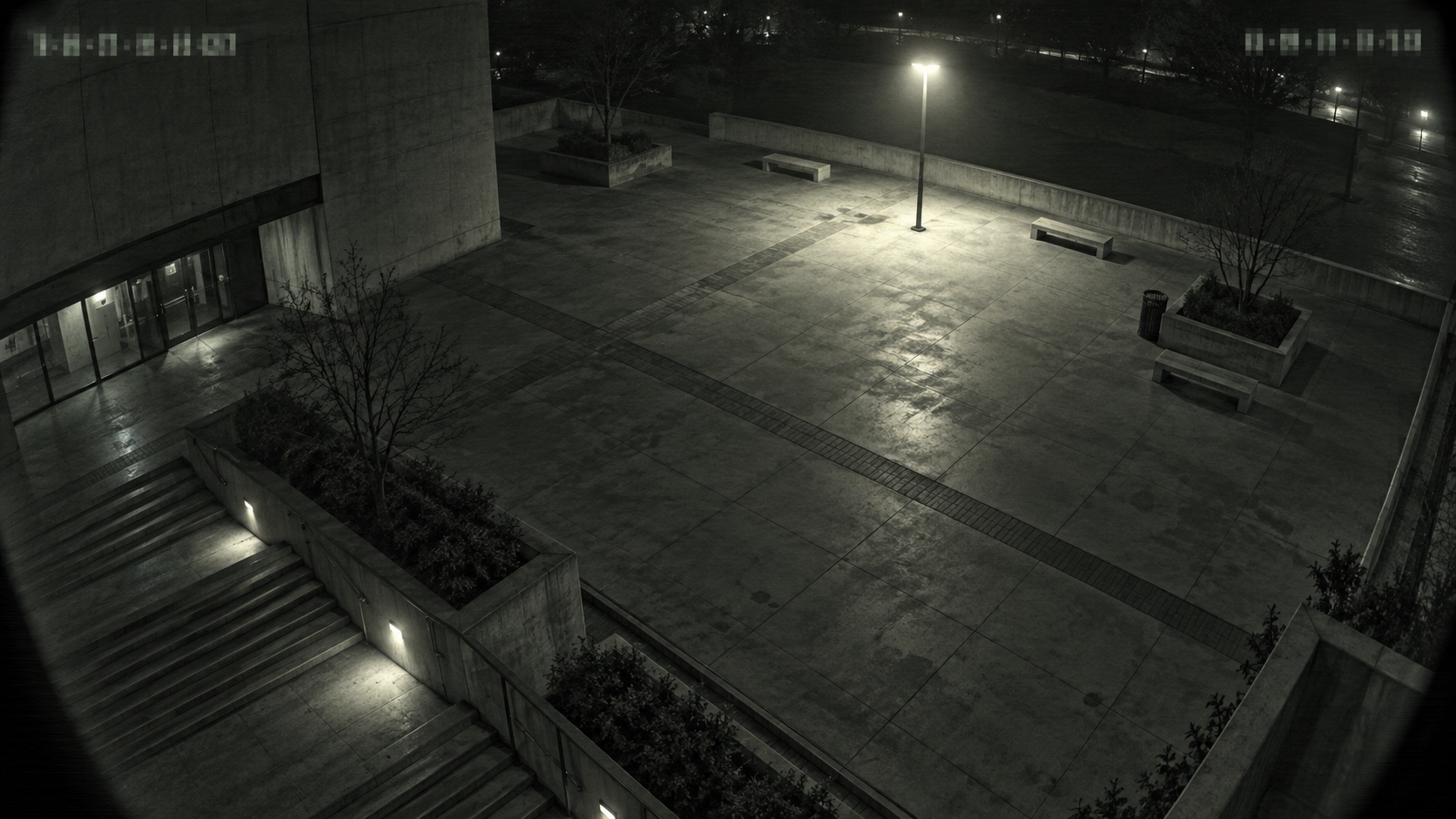 A deserted plaza at night under one lamp, shot from above in grainy monochrome.